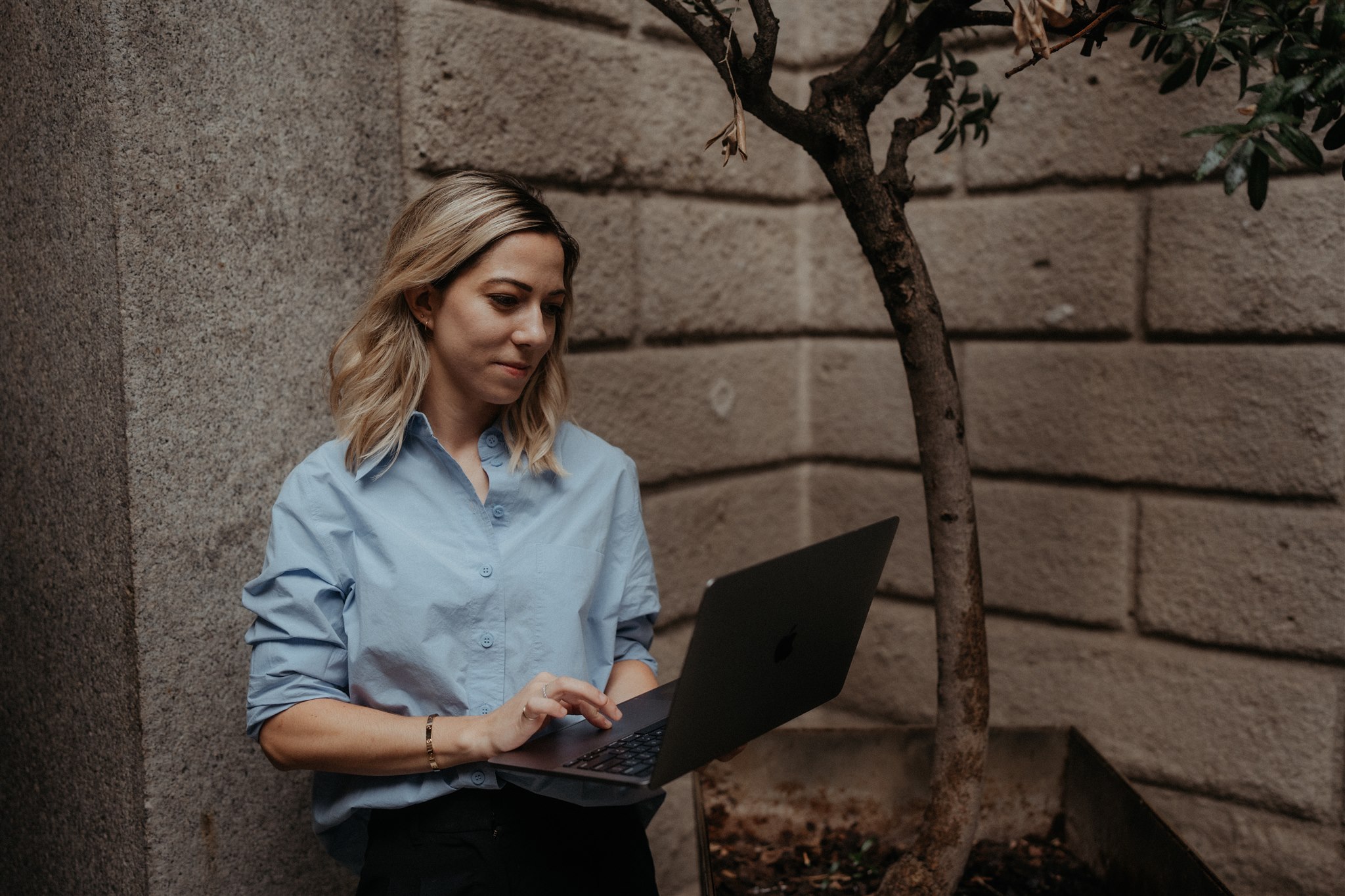 Woman leans against a concrete wall while holding her laptop and scrolling.