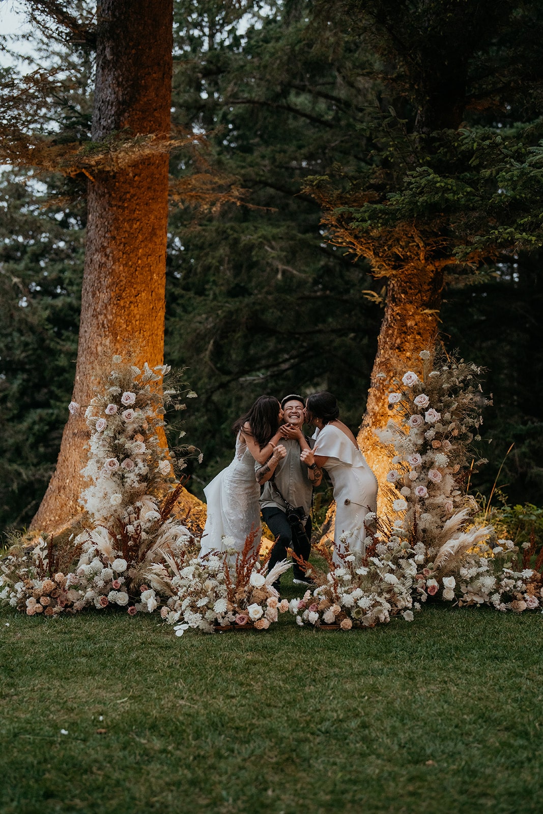 Two brides kiss their wedding photographer on the cheek during their micro wedding in Oregon. 