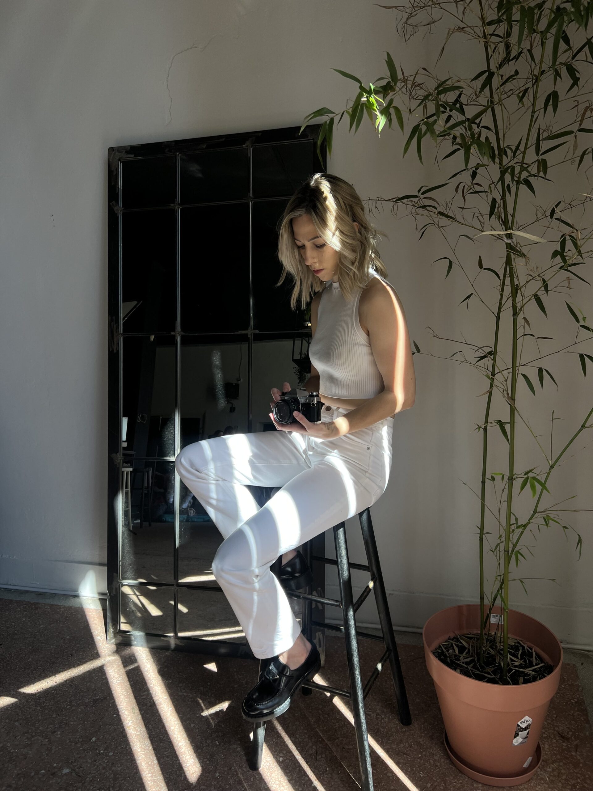 Female dressed in all white sits on a stool in a studio, adjusting her film camera settings.