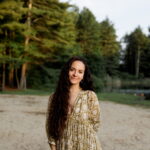 A female East Coast wedding photographer stands on a sandy beach with a forest in the background.