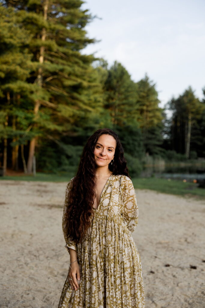 A female East Coast wedding photographer stands on a sandy beach with a forest in the background.